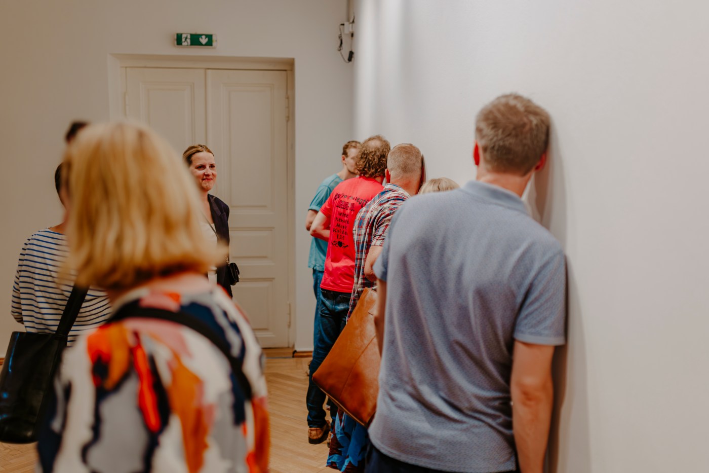Visitors stand with their ears pressed against the white wall, listening.