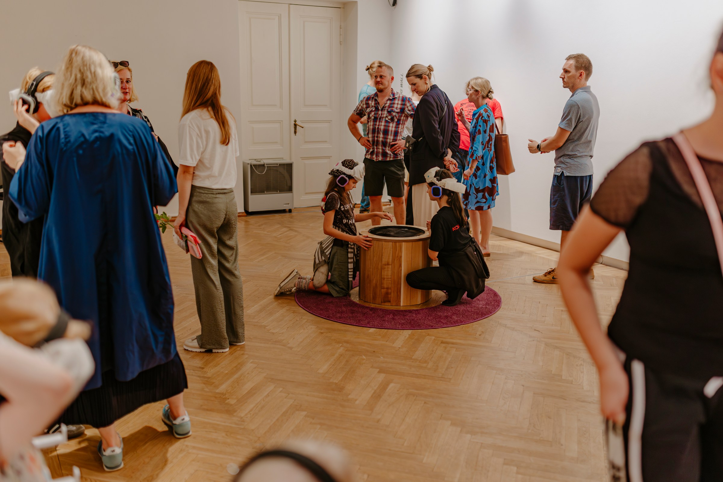 Families gather around the well in the gallery; children with headphones kneel at the water.