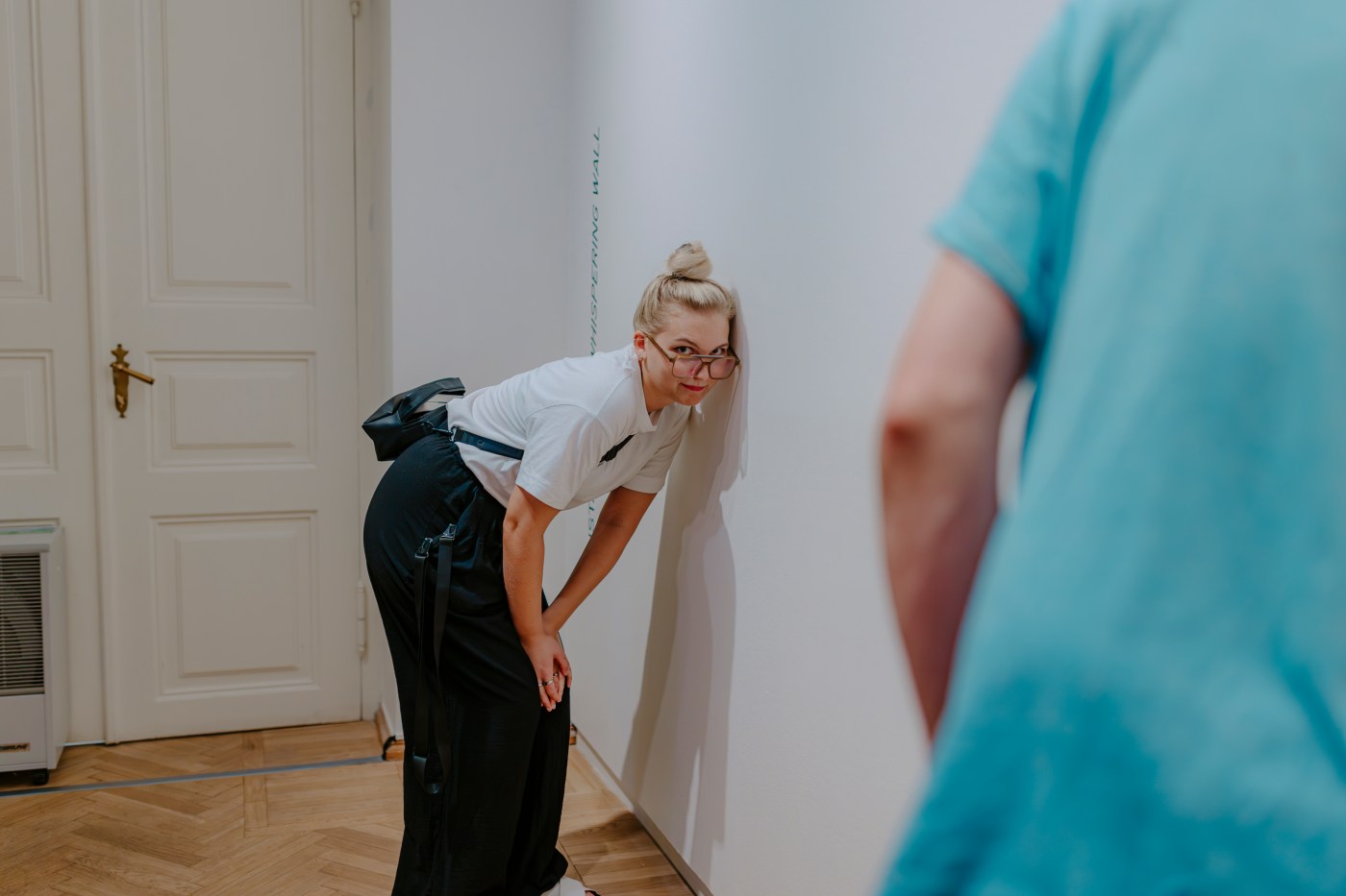 A woman bends forward and puts her ear to the wall.