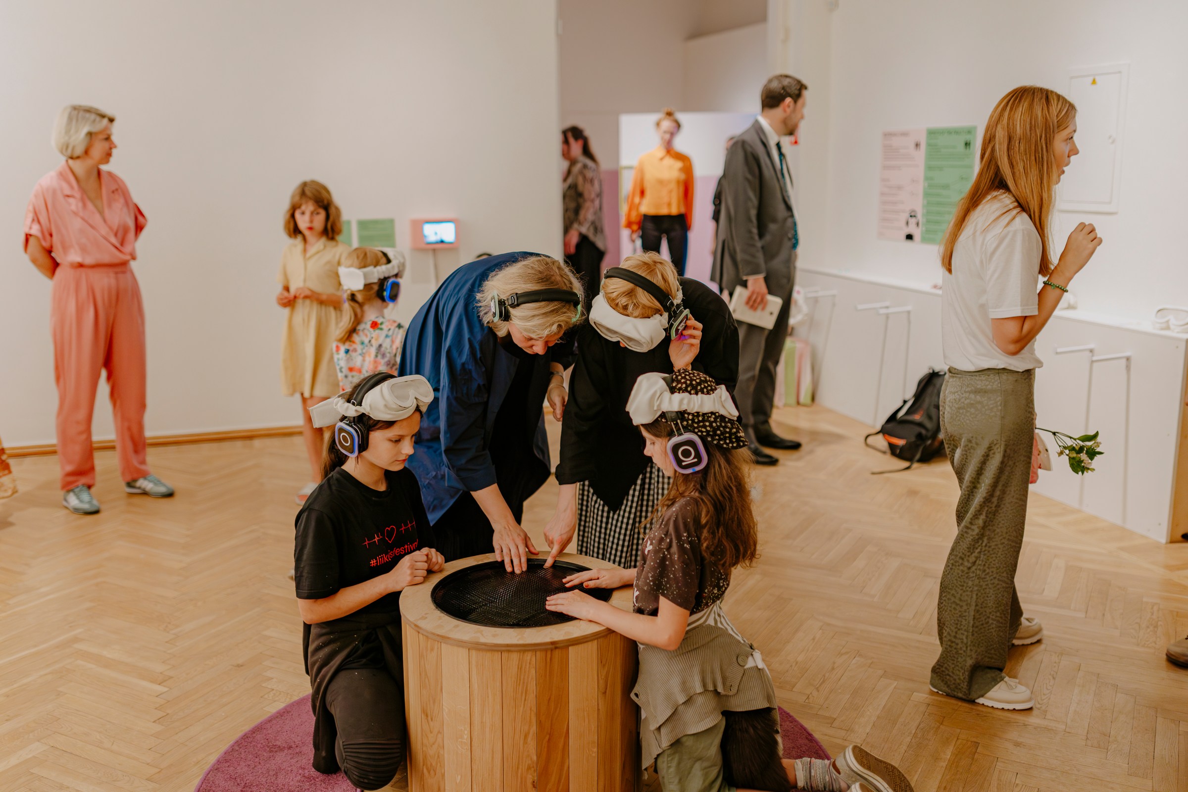 Children kneel around the wooden well with headphones on, touching the black water while adults watch.