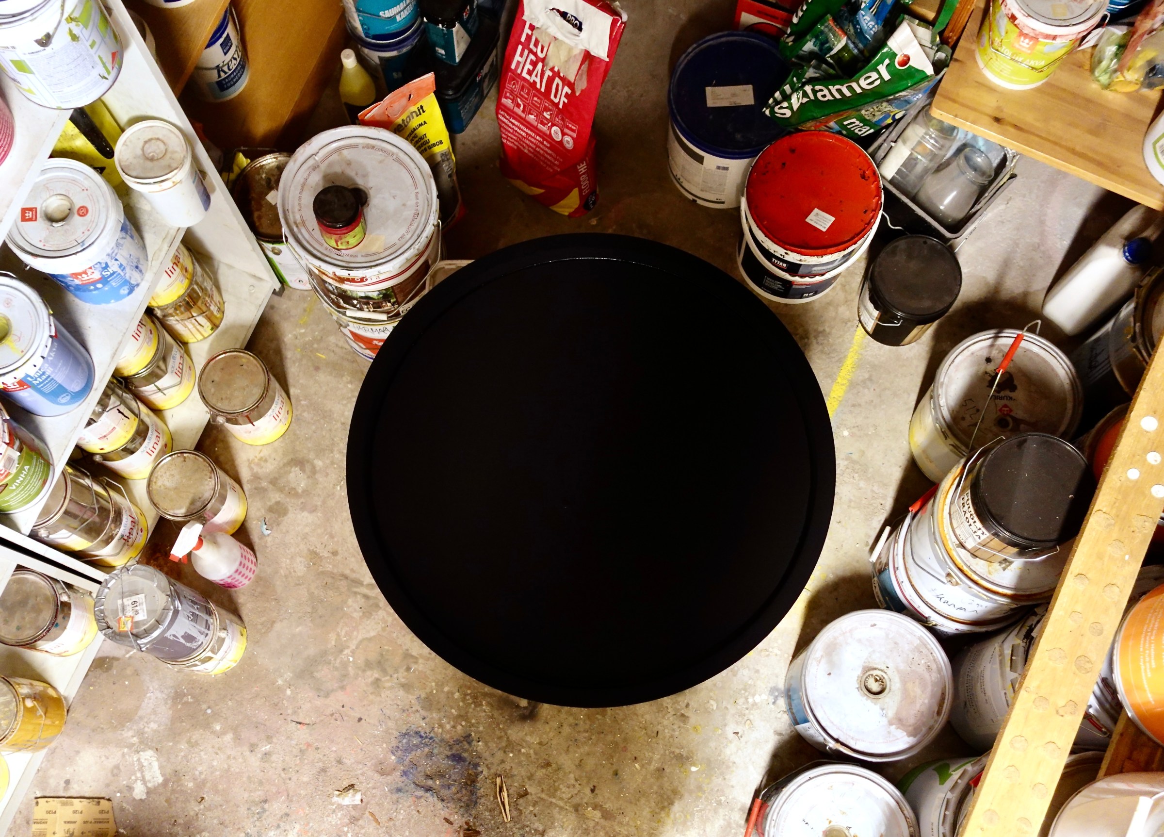 A water basin painted with Mussou Black seen from above, among paint tins in the studio. The black surface reads as a flat hole.