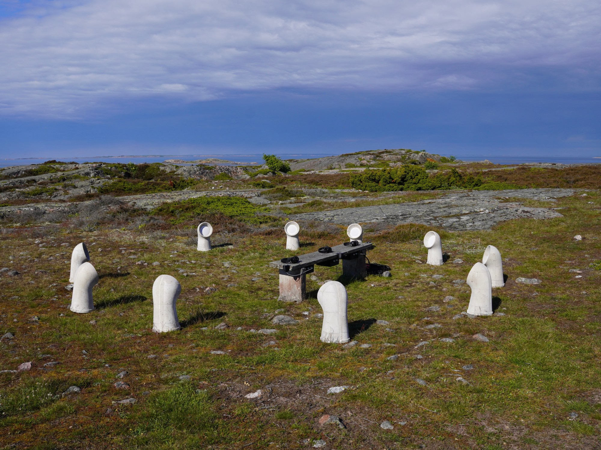 A circle of hand-cast concrete cowl speakers placed low among rock and juniper on Jurmo island, sea and sky in the distance