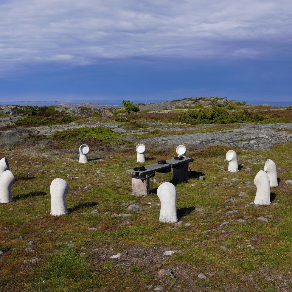 A circle of hand-cast concrete cowl speakers placed low among rock and juniper on Jurmo island, sea and sky in the distance