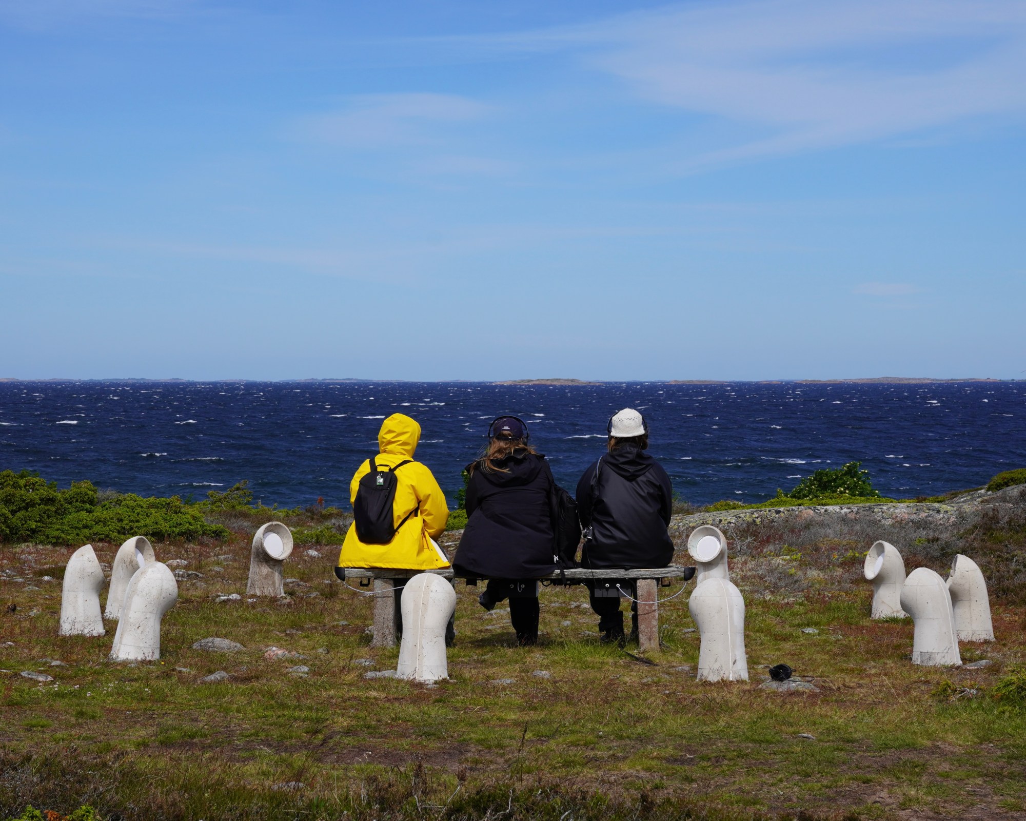 Three visitors seen from behind, sitting on the bench and listening toward the Baltic Sea.
