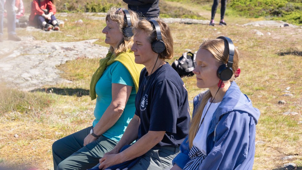 Visitors sitting on a bench and listening to the INTER, Jurmo installation through headphones near Jurmo chapel and the Baltic Sea. Visitors listening to INTER, Jurmo during the opening event. Photo: Joonas T. Laine.