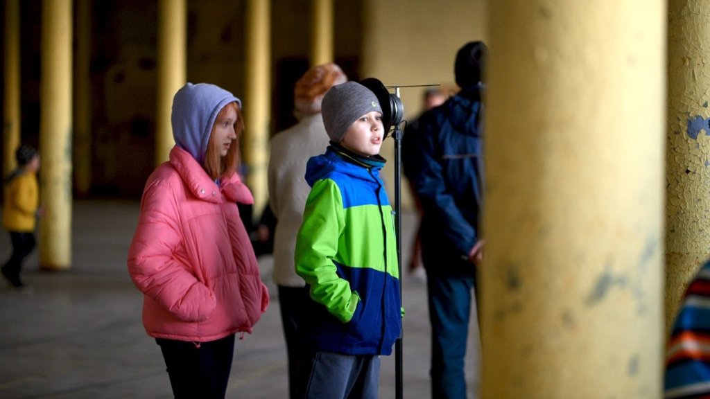 Two children standing beside a loudspeaker among the columns, listening.
