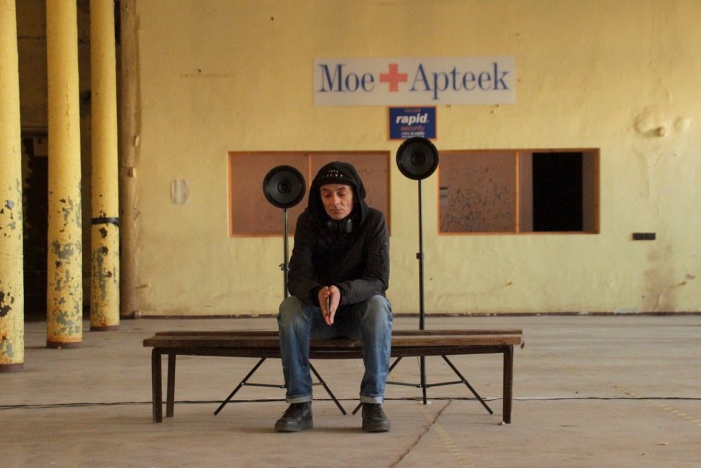 A man sitting alone on a bench between two loudspeakers under an old Moe Apteek sign.