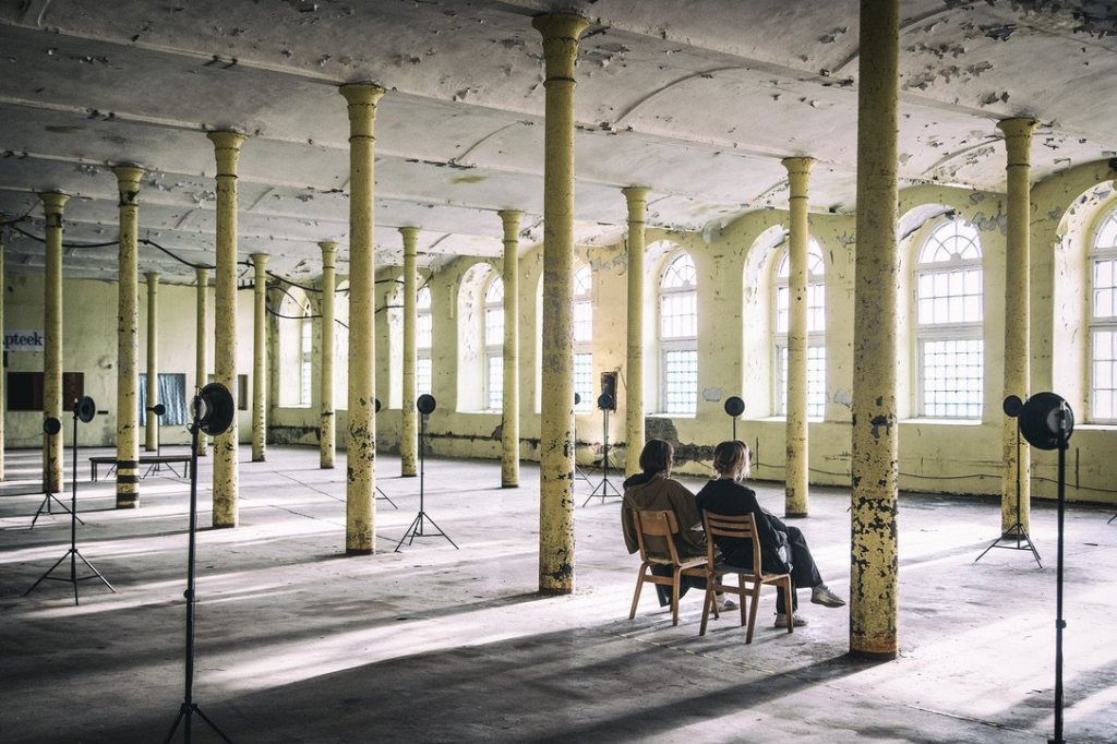 Two people sitting on chairs in the sunlit factory hall, surrounded by loudspeakers on stands.
