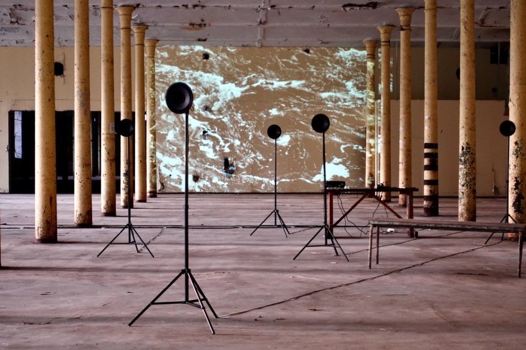 Loudspeakers on stands in front of a wall-sized projection of moving river water.