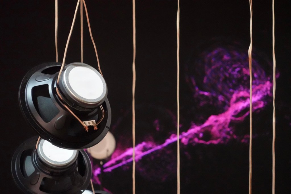 Bare loudspeaker drivers hanging on cables in front of the purple generative image NIMITTA.