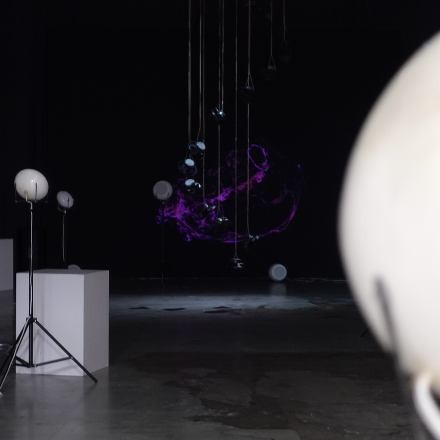 View past a blurred white breath speaker towards the suspended speaker column and the purple projection.