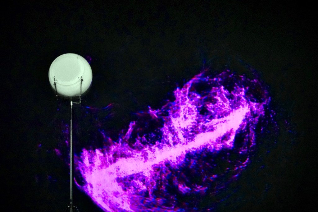A white breath speaker on a stand beside the purple generative image NIMITTA on a black wall.