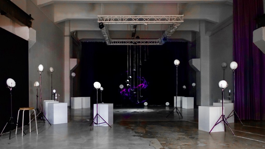 RE-BIRTH in the lit hall at MUU Helsinki: white breath speakers on stands and plinths lead towards the suspended speaker column and projection.