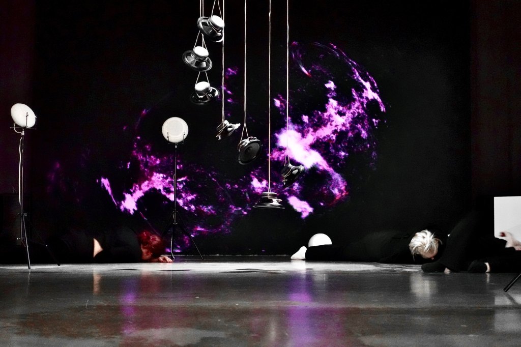 People lying on the floor beneath suspended speakers, with NIMITTA on the dark wall behind them.