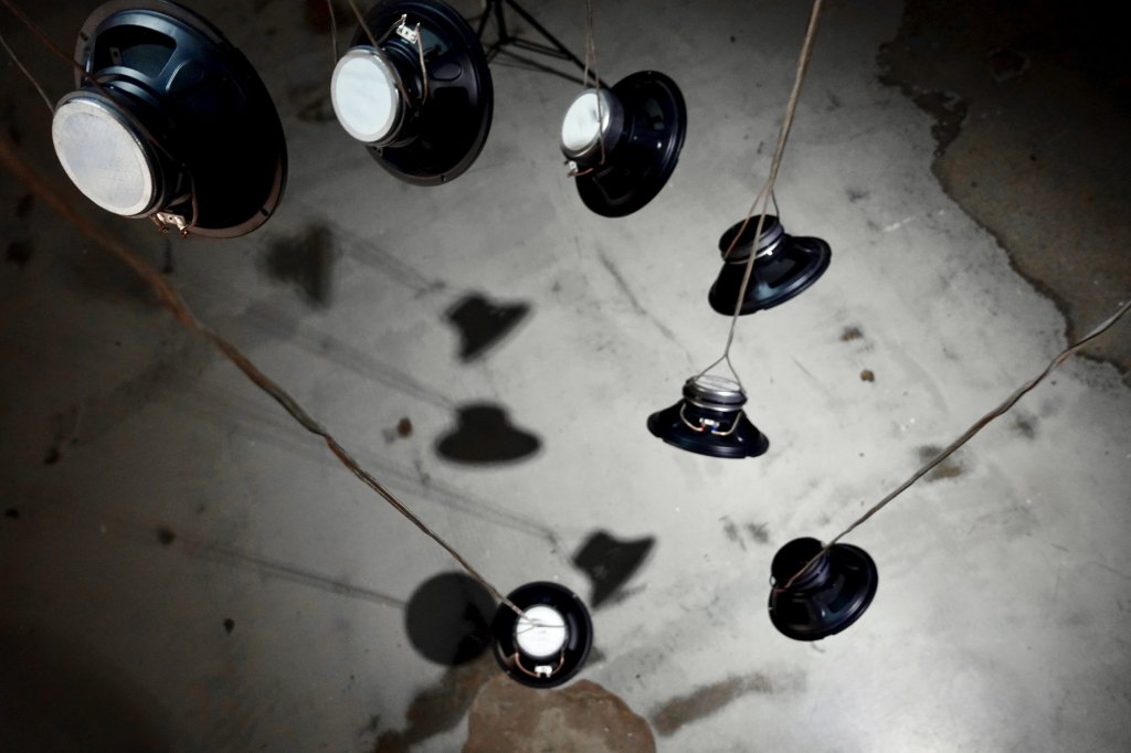 Bare loudspeaker drivers hanging on cables, their shadows on a concrete floor.